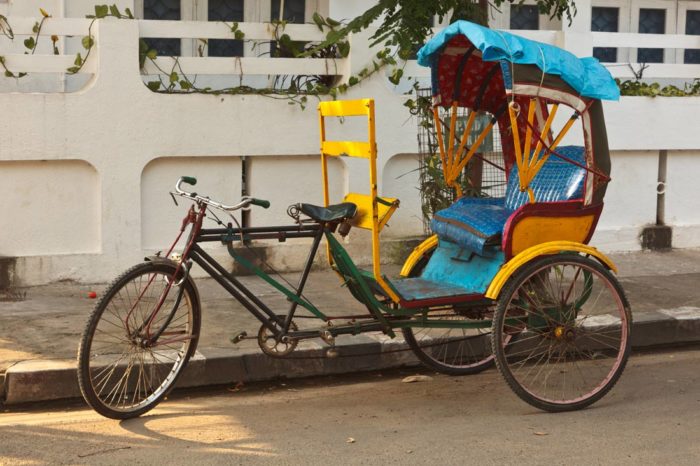 A colorful three-wheeled pedal rickshaw with a blue canopy parked beside a white wall under warm daylight.
