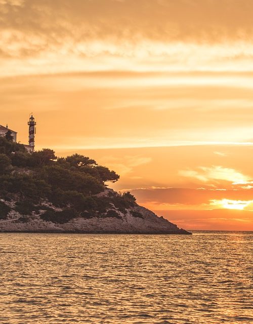 A rocky, tree-covered point with a lighthouse set against an orange and gold sunset sky over the sea.