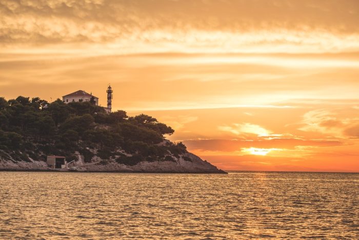 A coastal headland with a lighthouse and trees silhouetted against a glowing orange sunset over calm water.